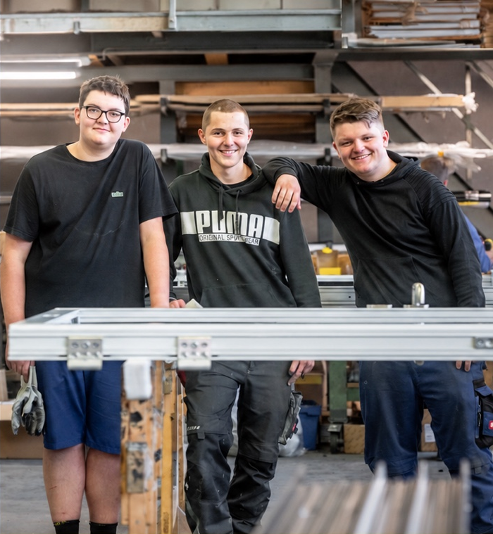 Drei junge Männer aus dem Saller-Team lächeln in der Werkstatt hinter einem Metallrahmen